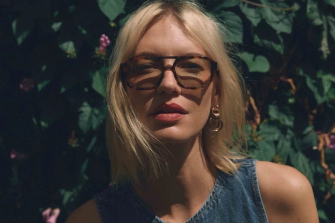 Blonde woman wearing tortoiseshell aviator glasses and a denim top against a leafy green garden background.