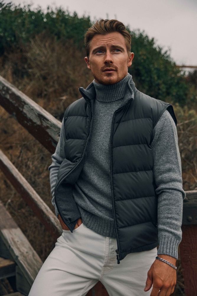 Man in a puffer vest and turtleneck sweater stands on wooden steps outdoors, showcasing fashion and lifestyle in overcast weather.