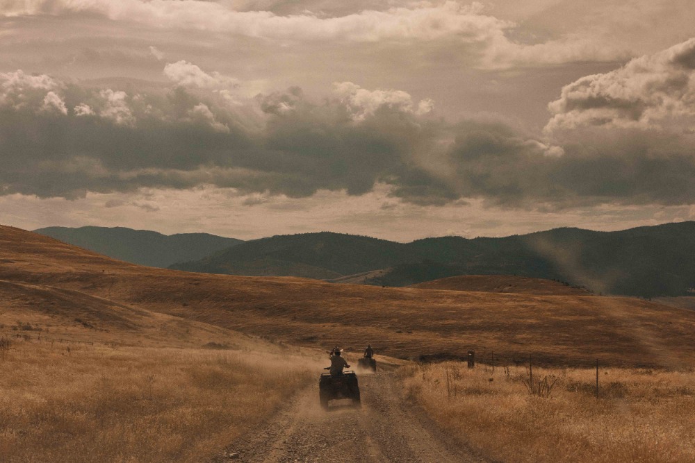 Two people ride ATVs on a dirt road through a golden field, under a cloudy sky, with mountains in the distance, conveying outdoor recreation.