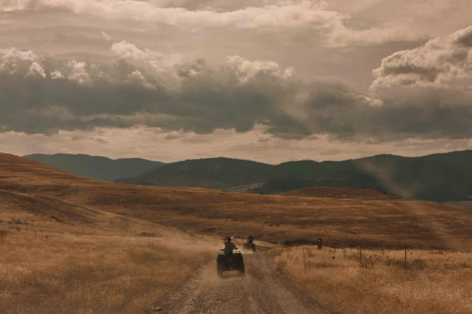 Two people ride ATVs on a dirt road through a golden field, under a cloudy sky, with mountains in the distance, conveying outdoor recreation.
