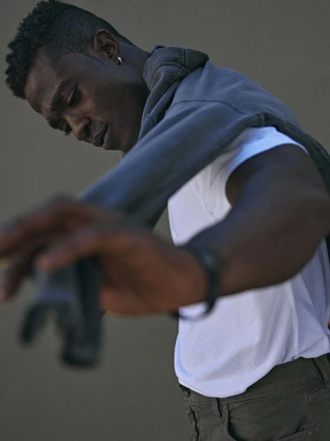 Young Black man in casual fashion leaning forward wearing a white tee, olive pants, a grey hoodie, and silver hoop earring.