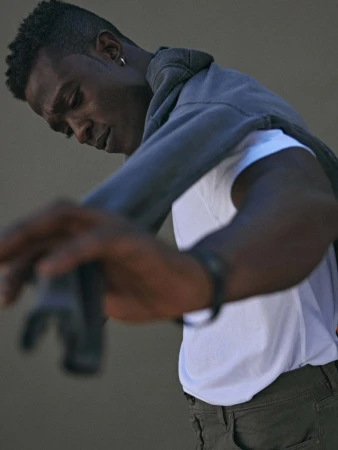 Young Black man in casual fashion leaning forward wearing a white tee, olive pants, a grey hoodie, and silver hoop earring.
