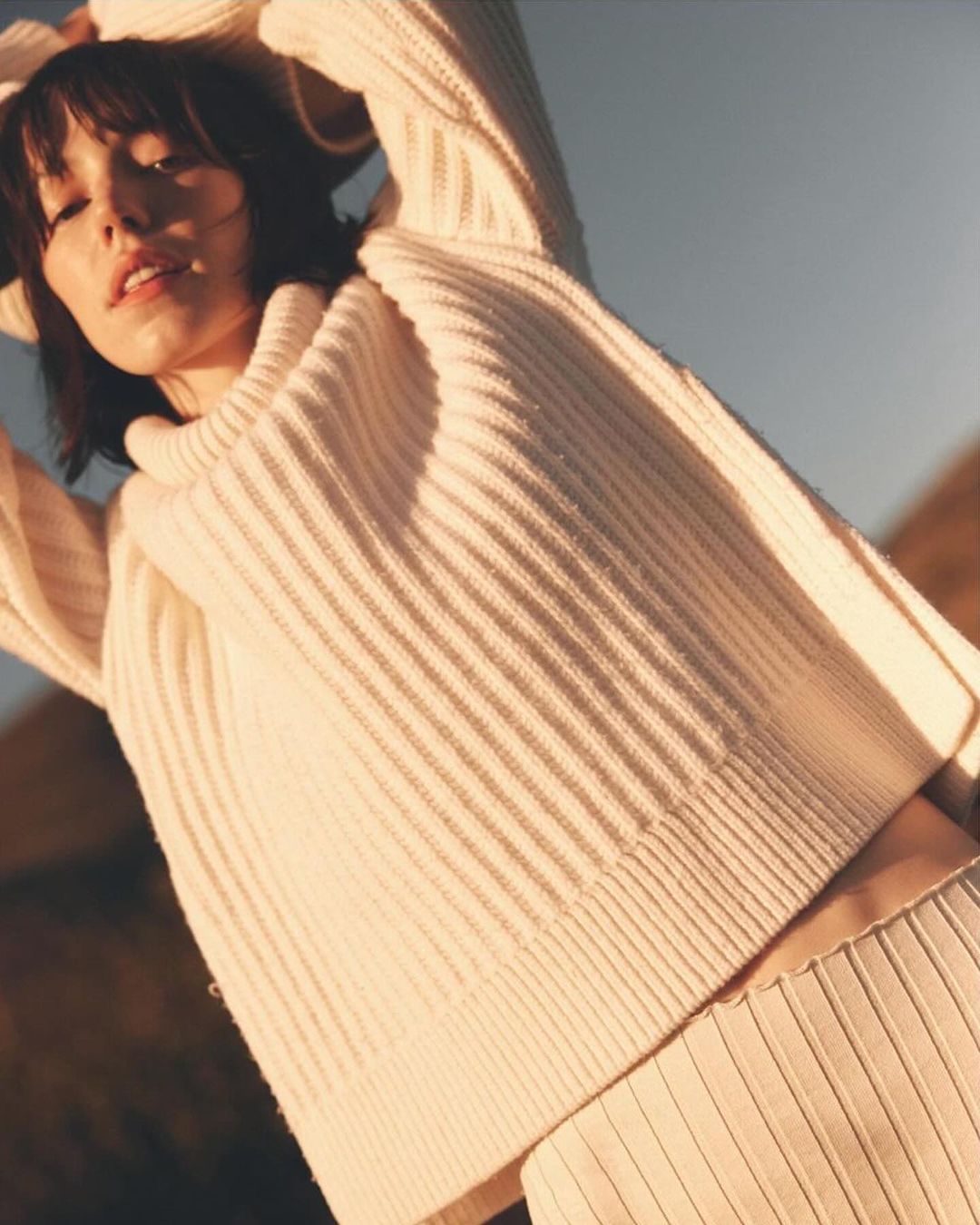 Woman in a cream sweater and skirt poses outdoors with her arms raised in a fashion shot with a natural background and soft lighting.