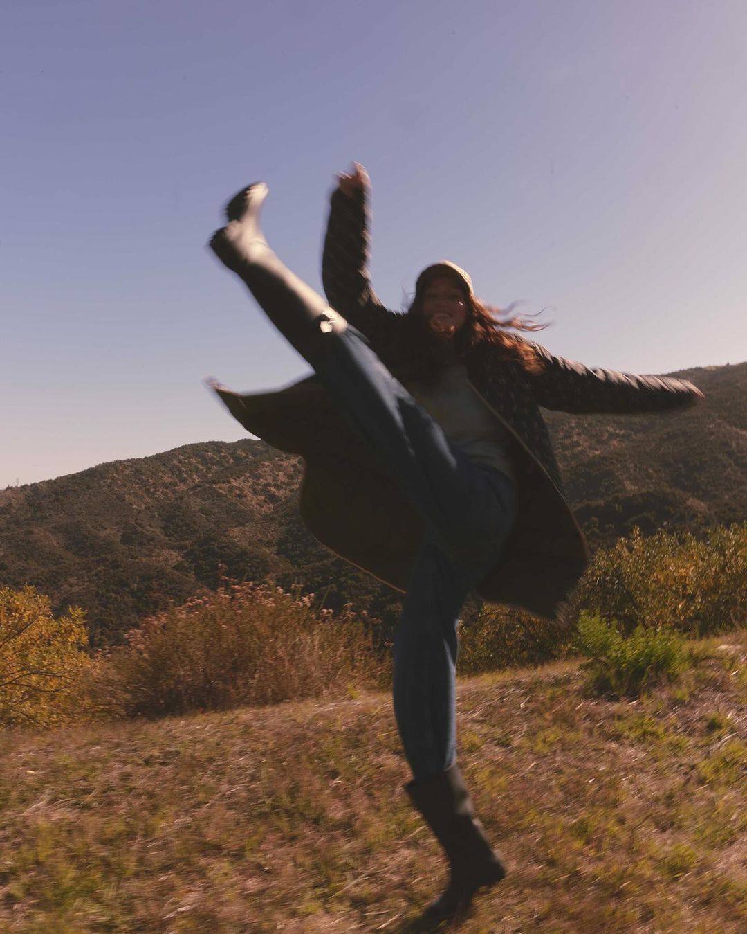 A woman in jeans, boots, and a coat kicks her leg high in the air on a grassy hillside under a clear blue sky with soft focus.