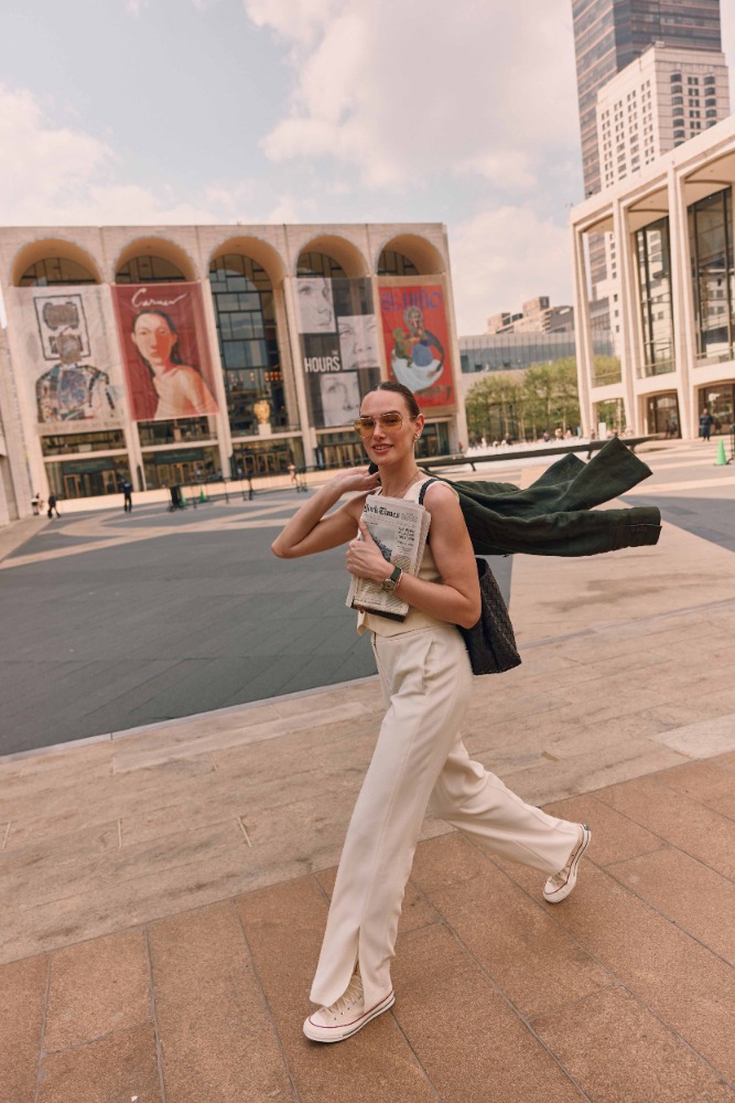 Woman walks in front of the David Koch Theater in NYC, holding a newspaper, wearing sunglasses, cream-colored pants, and a tank top.