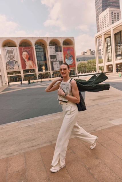 Woman walks in front of the David Koch Theater in NYC, holding a newspaper, wearing sunglasses, cream-colored pants, and a tank top.