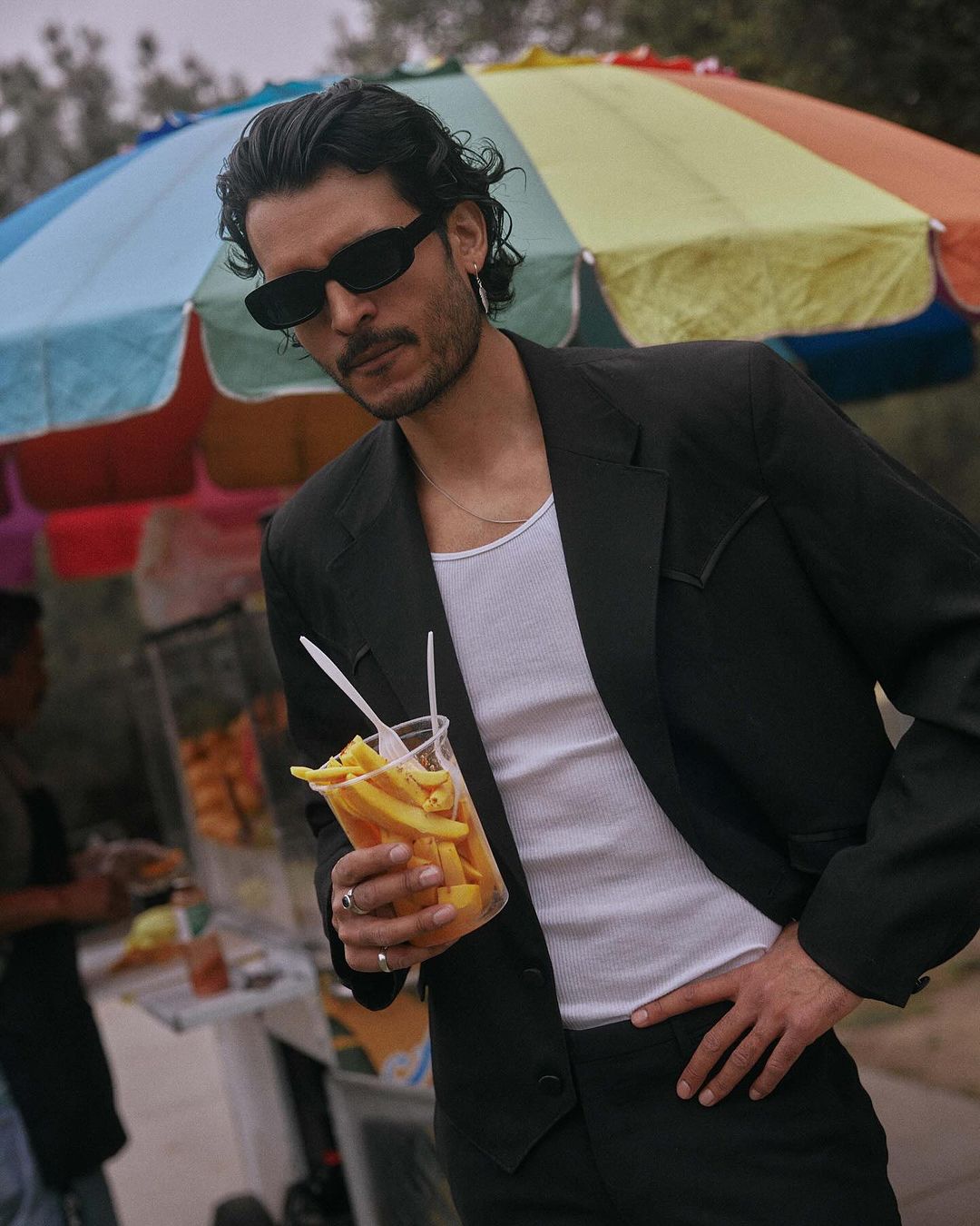 Portrait of a stylish adult man wearing sunglasses and a black suit, holding a cup of fries near a food cart outdoors.