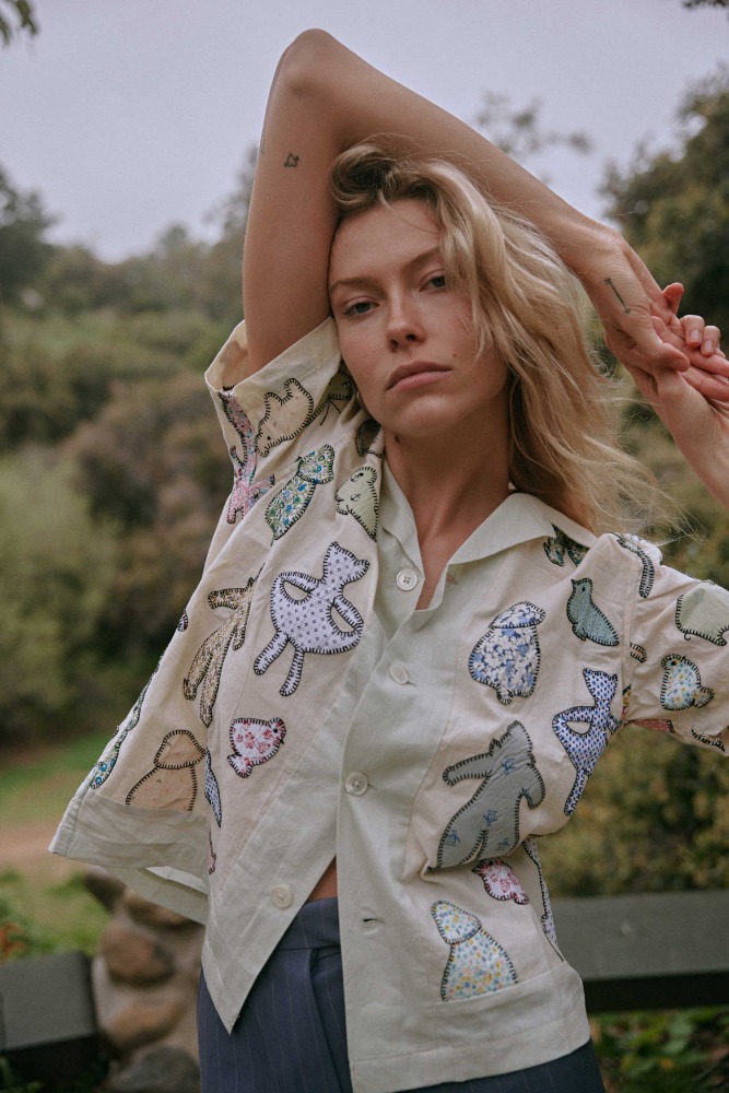 Blonde woman in embroidered button-down shirt poses against a green leafy background, fashionably styled, shot in natural lighting.