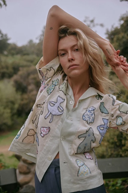 Blonde woman in embroidered button-down shirt poses against a green leafy background, fashionably styled, shot in natural lighting.