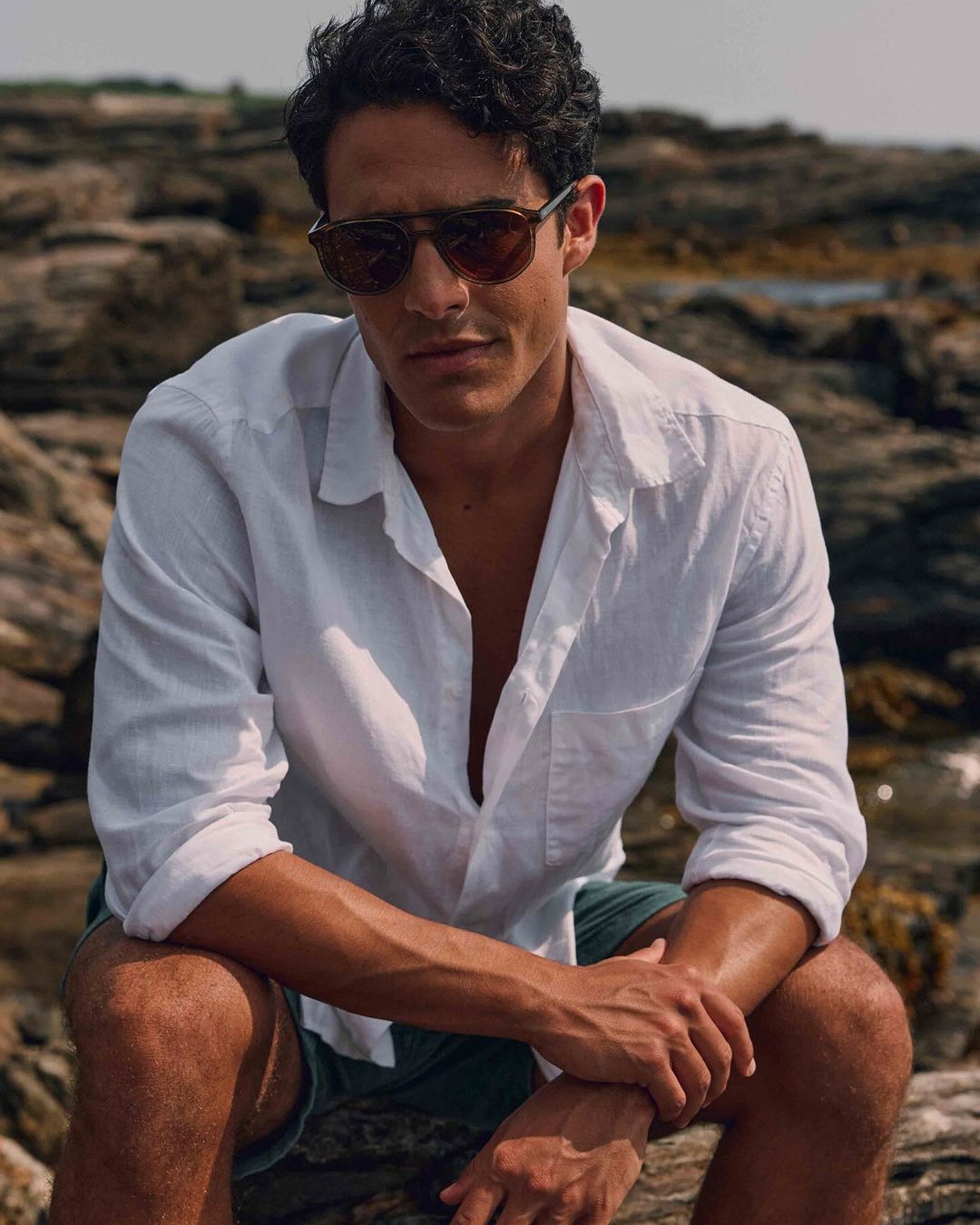 Portrait of a man in a white button-down shirt and sunglasses sitting on rocks by the ocean, showcasing natural style and coastal setting.