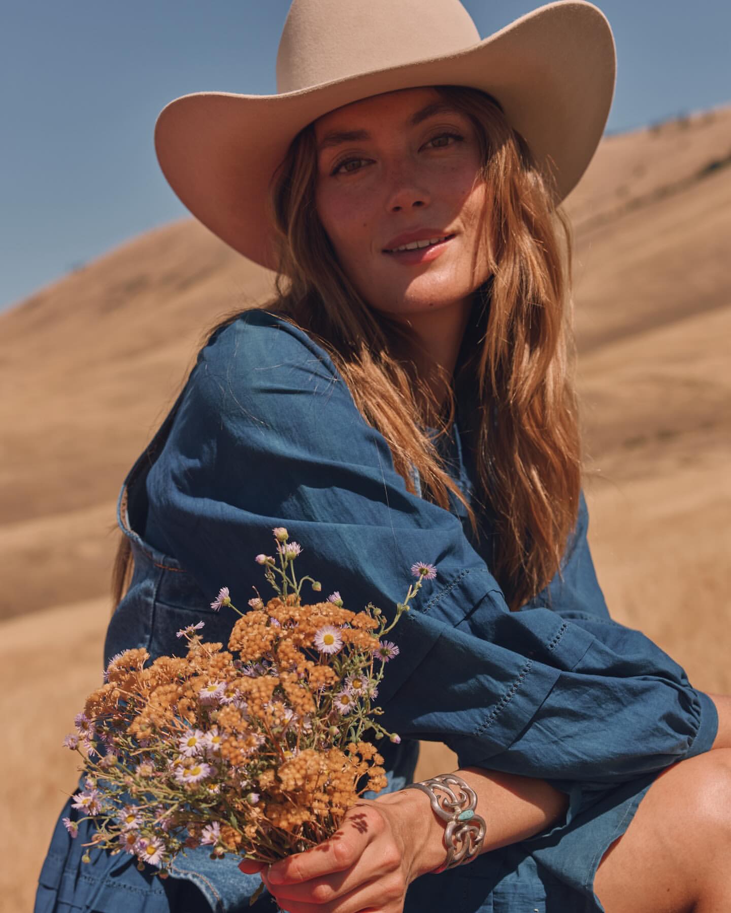 Woman wearing a wide-brimmed hat and blue dress holding a small bouquet of wildflowers in a dry landscape by John Schell.