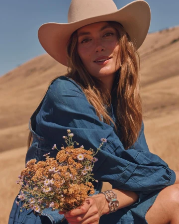 Woman wearing a wide-brimmed hat and blue dress holding a small bouquet of wildflowers in a dry landscape by John Schell.