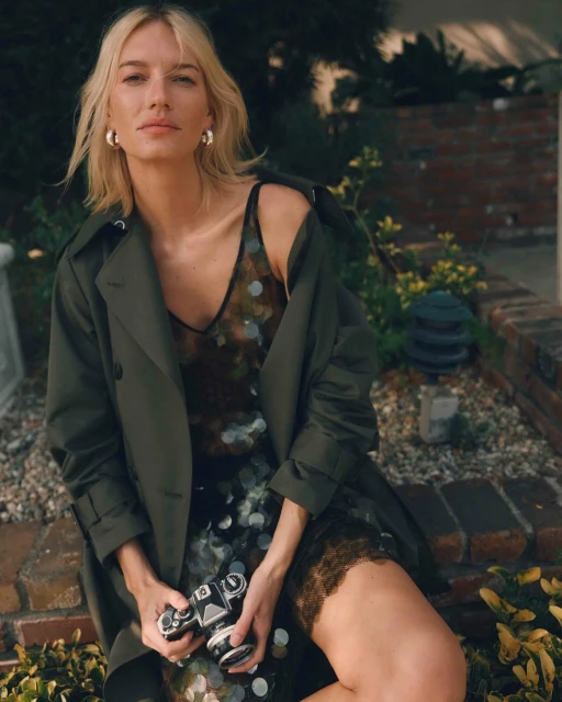 A blonde woman sits outside with a vintage camera, wearing a green coat over a patterned dress, posing on a brick wall in daylight.