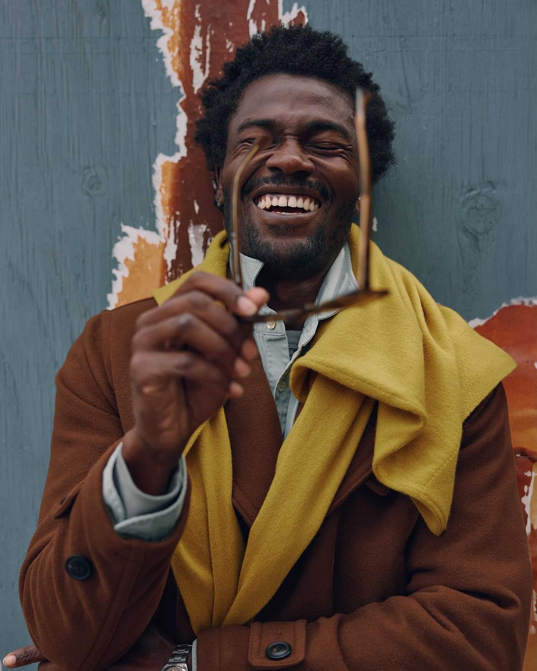 Happy Black male model in brown coat and mustard scarf holds eyeglasses, posing against aged blue wall for fashion or lifestyle editorial.