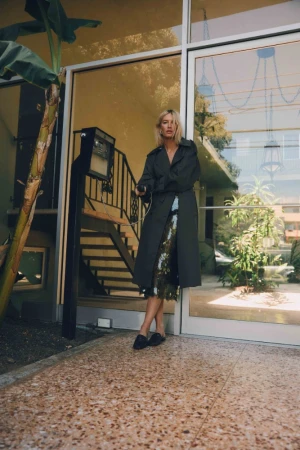 Blonde woman wearing a long dark trench coat standing before glass doors of a residential building in Los Angeles.