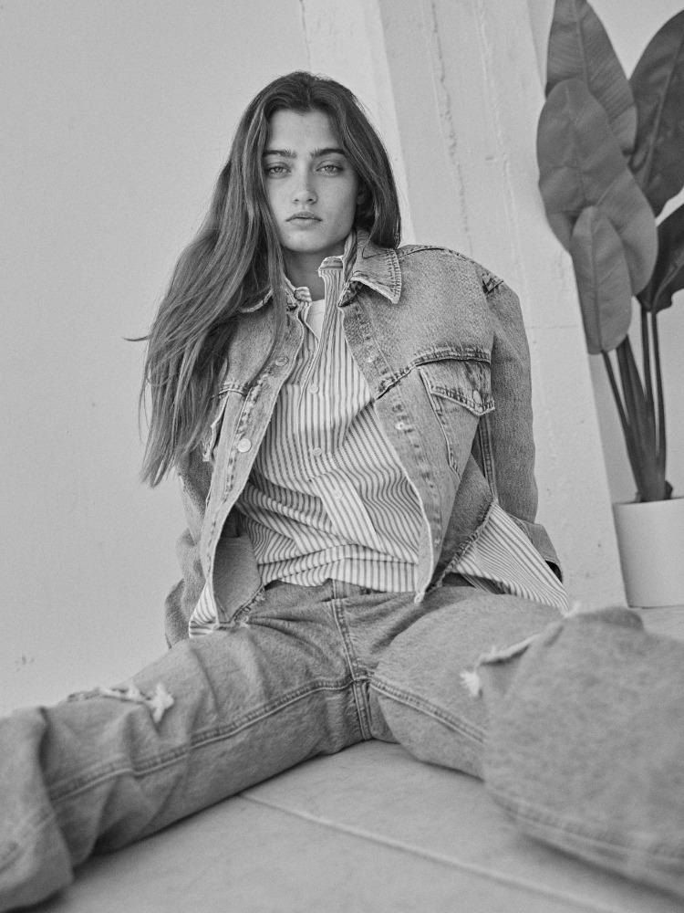 Black and white portrait of a young woman with long hair, wearing a denim jacket and jeans, sitting indoors with plants in background.