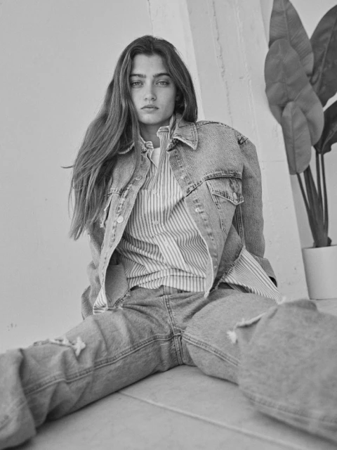 Black and white portrait of a young woman with long hair, wearing a denim jacket and jeans, sitting indoors with plants in background.