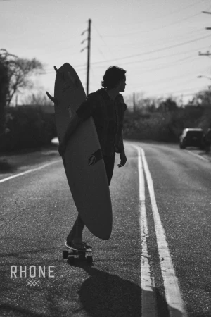 Monochrome photograph of a surfer riding a skateboard down an asphalt road holding a surfboard under his arm.