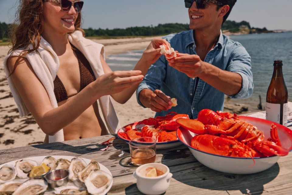 A woman and man enjoy lobster, oysters, and drinks at a picnic table on a sunny beach, with water and greenery in the background.