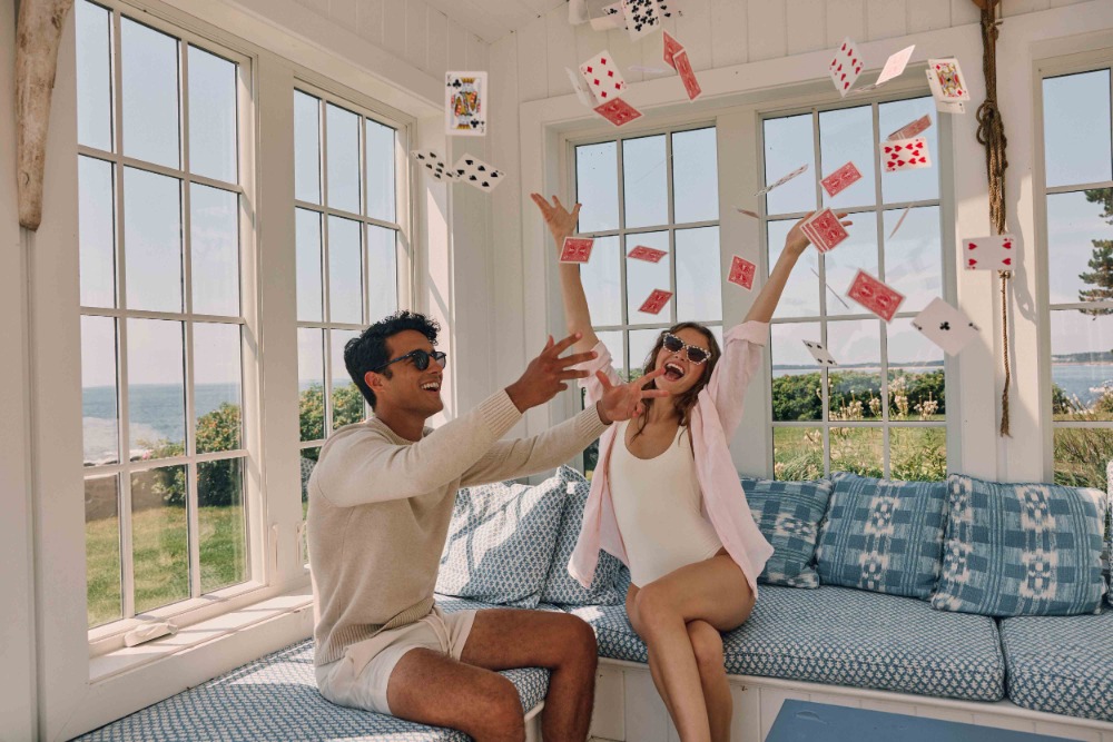 Two people laugh as playing cards fly near windows overlooking the ocean, sitting on a built-in blue-patterned cushioned bench.