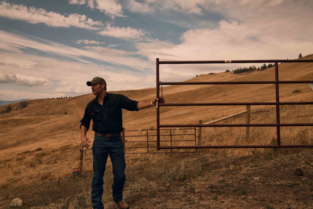 Man standing near metal gate in rural landscape with rolling hills and cloudy sky in natural light, wearing a baseball cap and dark clothing.
