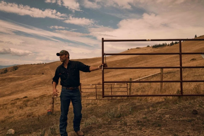 Man standing near metal gate in rural landscape with rolling hills and cloudy sky in natural light, wearing a baseball cap and dark clothing.