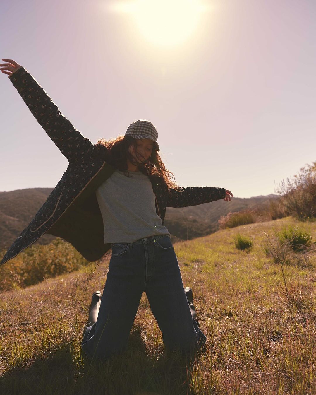 Woman in outdoor setting wearing coat and jeans, with arms outstretched towards the sun on grass hillside with mountains in background.