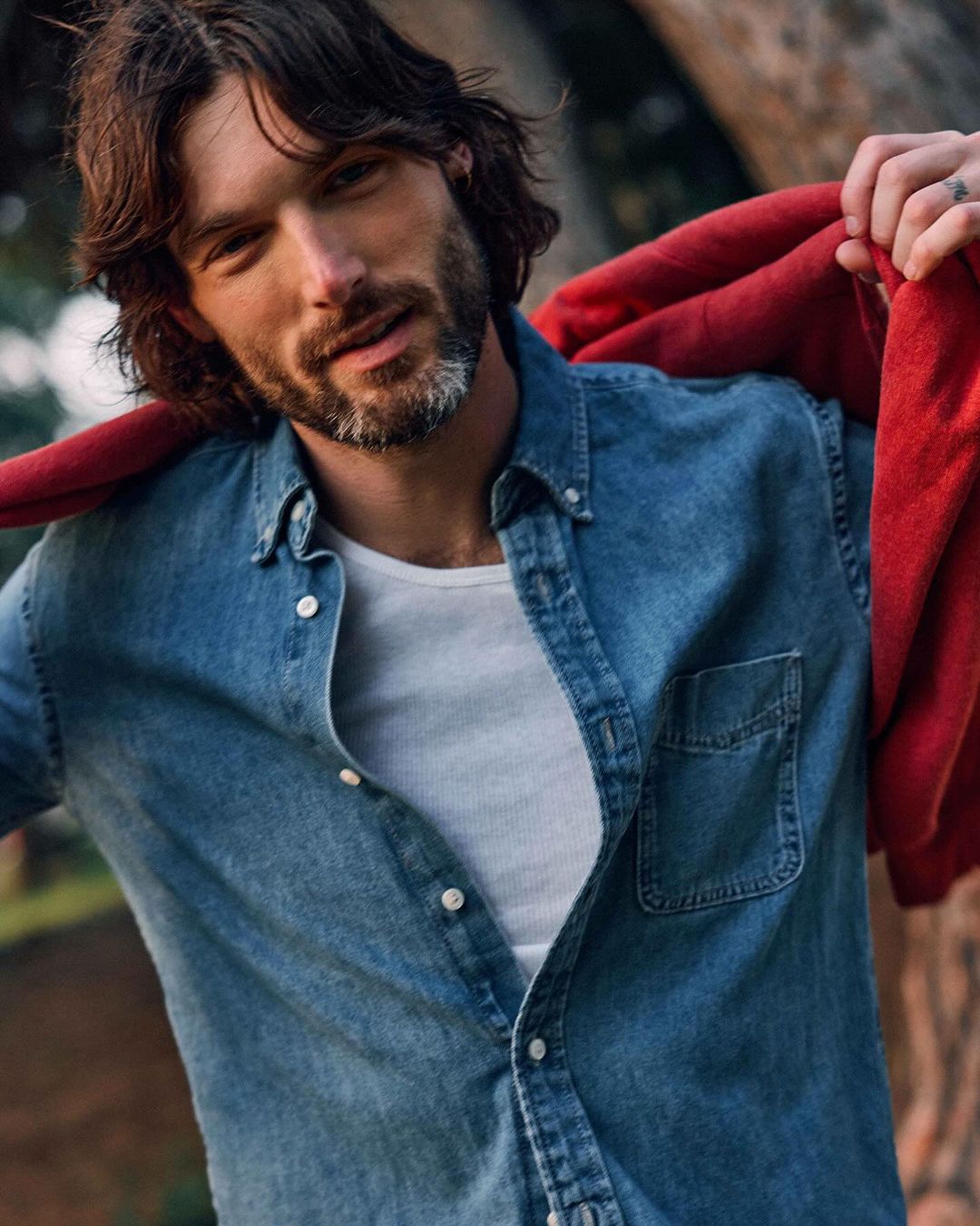 Portrait of a brown-haired man with a beard in a blue denim shirt and white t-shirt outdoors, wearing a red jacket over his shoulder.