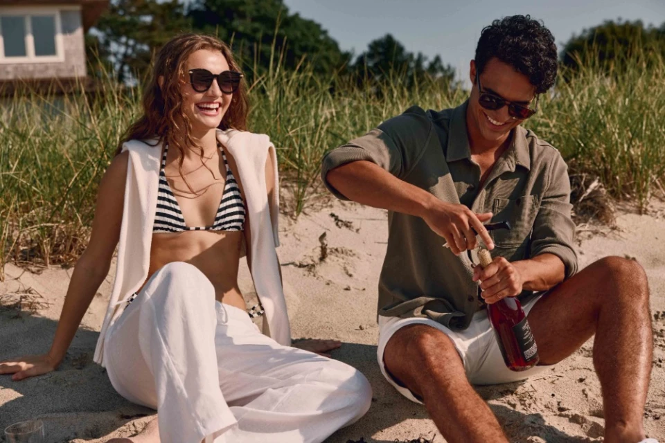 A smiling man and woman sit on a sandy beach. The man opens a bottle of rose wine, with dune grass and a house in the background.