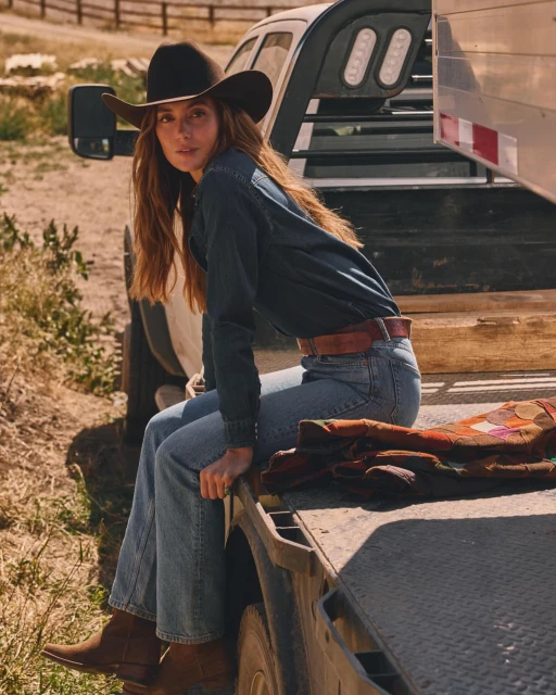 Woman in cowboy hat and denim outfit sits on a truck bed in an outdoor setting, creating a Western-themed fashion shot with natural light.