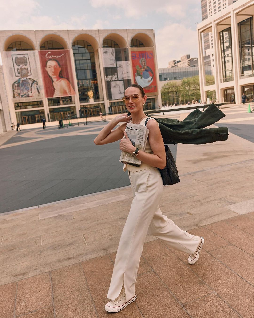 Stylish adult woman walks in front of theater in beige pants and top, carrying newspaper and coat slung over her shoulder, on sunny day.