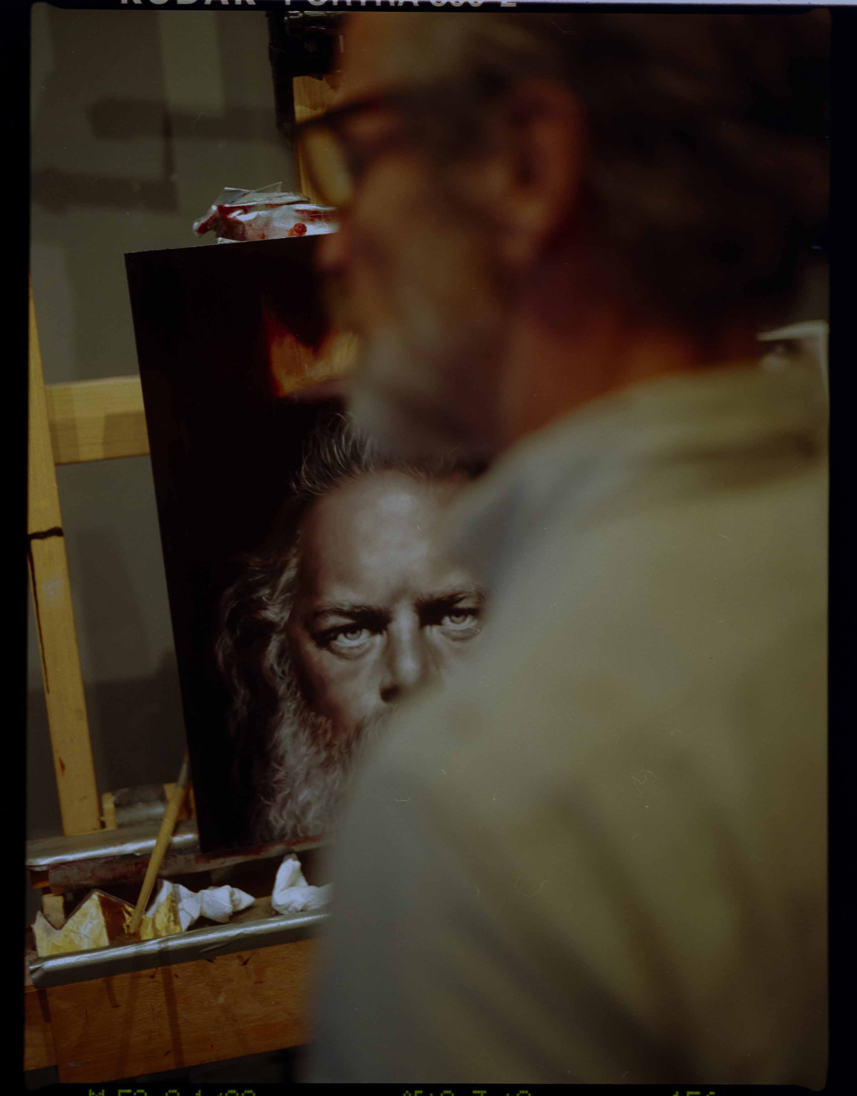 Portrait of a painter in art studio working on a painting of a man with beard. Painting on easel, art supplies, and glasses visible.