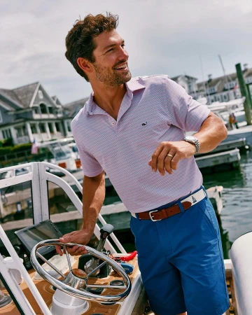 Smiling man in pink polo and blue shorts on a boat, houses in the background, daytime. Coastal fashion and summer sailing lifestyle.
