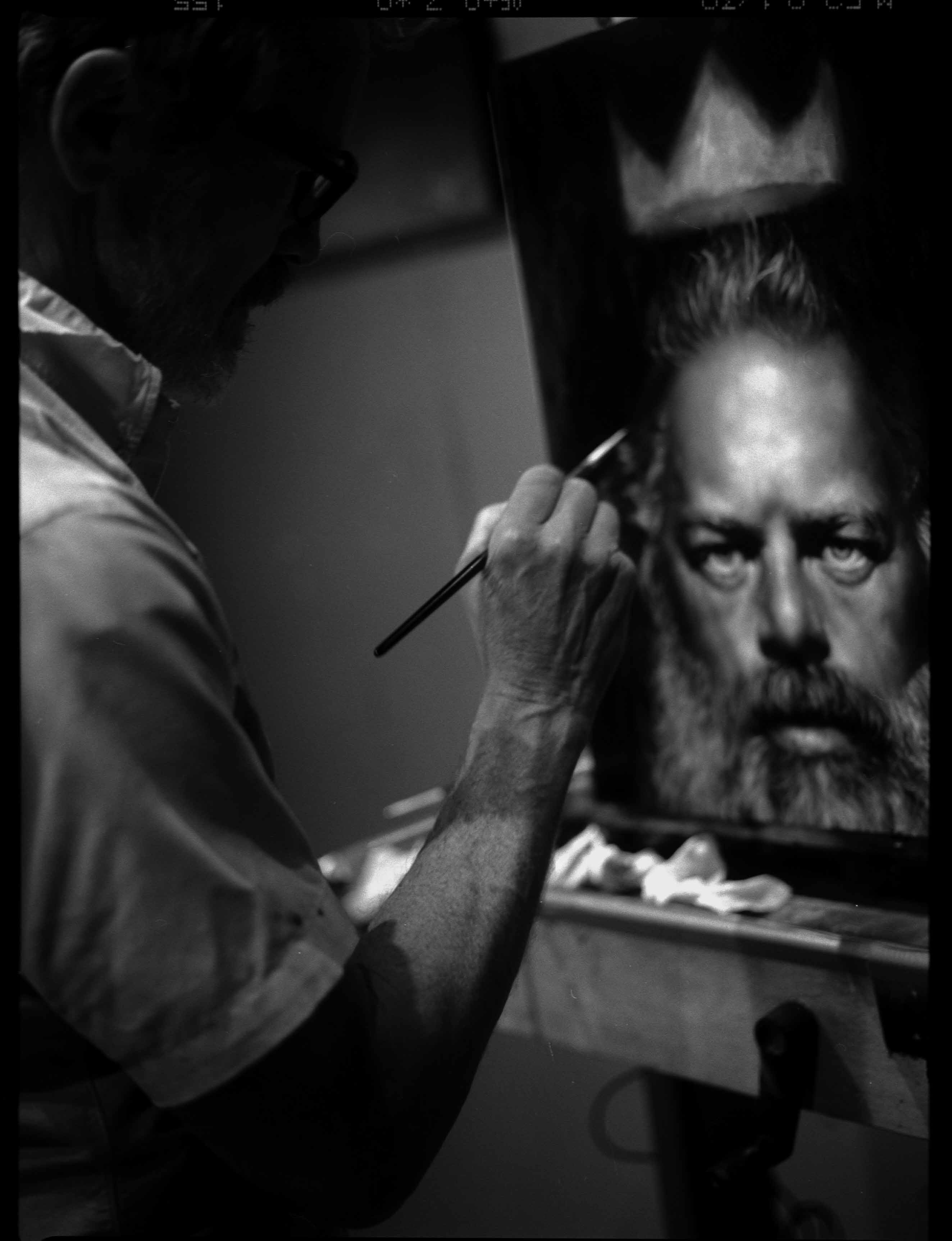 Black and white studio shot of a mature man painting a portrait of himself on a canvas, using a brush and standing indoors.