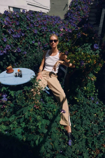 Adult female wearing a white tank top and khaki pants sitting outdoors in a lush garden setting with a blue table and drink.