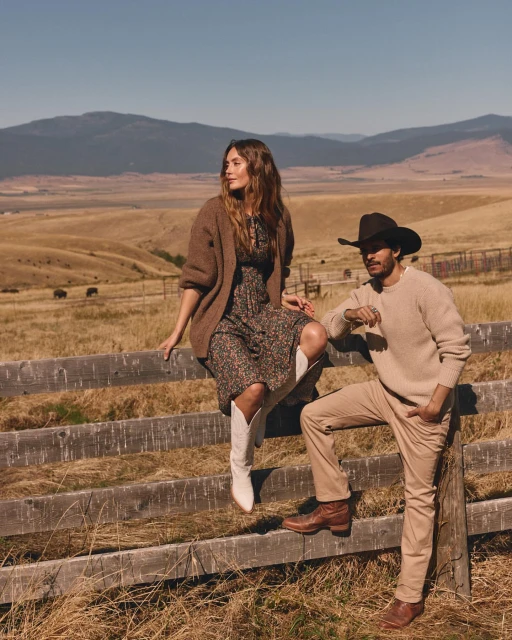 A casually dressed couple sits on a wooden fence in a grassy field with mountains in the background on a sunny day in the countryside.