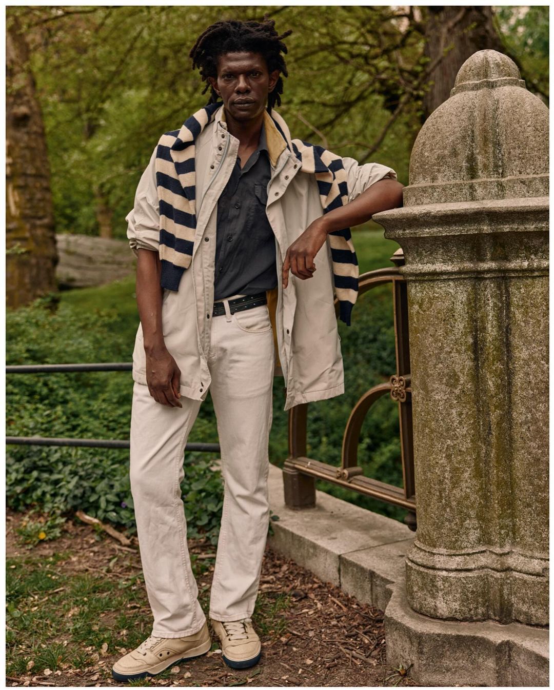 Adult male in a casual, layered outfit poses in an outdoor setting by a stone railing. He is wearing a jacket, shirt, and jeans.