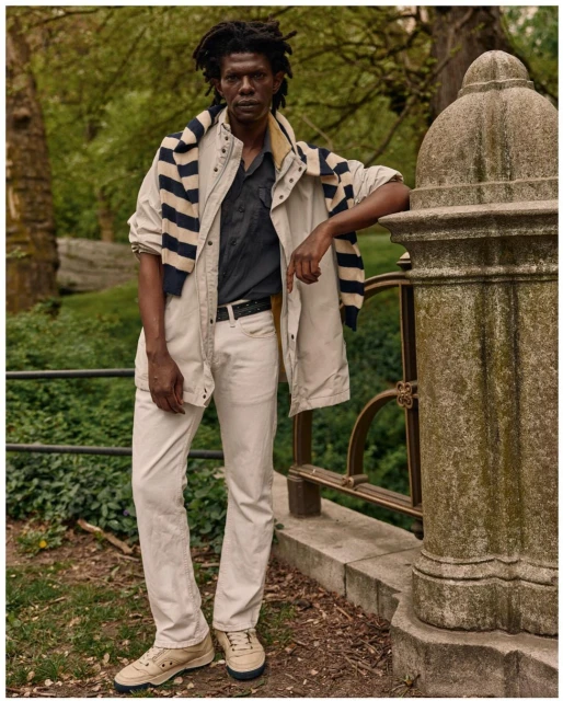 Adult male in a casual, layered outfit poses in an outdoor setting by a stone railing. He is wearing a jacket, shirt, and jeans.