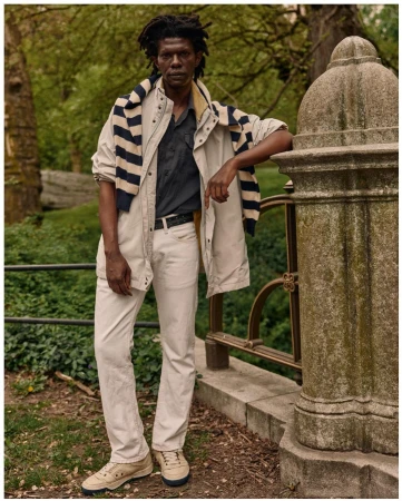Adult male in a casual, layered outfit poses in an outdoor setting by a stone railing. He is wearing a jacket, shirt, and jeans.