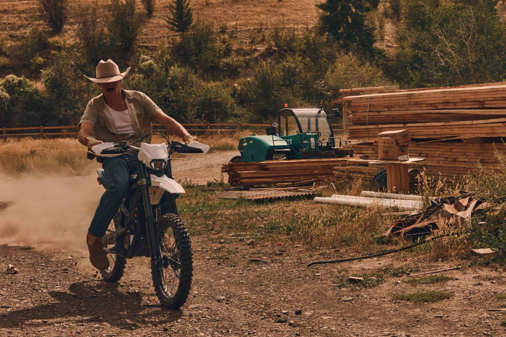 A man on a white dirt bike rides on a dirt road with an indistinct vehicle and logs behind him. He wears a cowboy hat and brown boots.