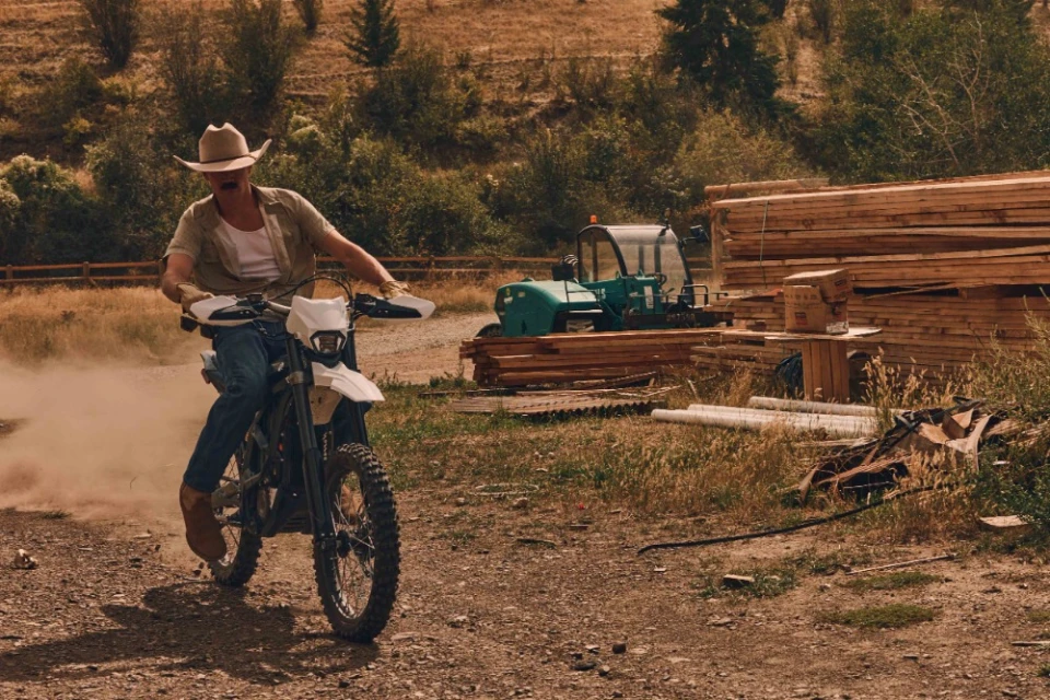 A man on a white dirt bike rides on a dirt road with an indistinct vehicle and logs behind him. He wears a cowboy hat and brown boots.