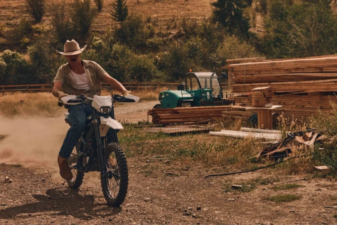 A man on a white dirt bike rides on a dirt road with an indistinct vehicle and logs behind him. He wears a cowboy hat and brown boots.