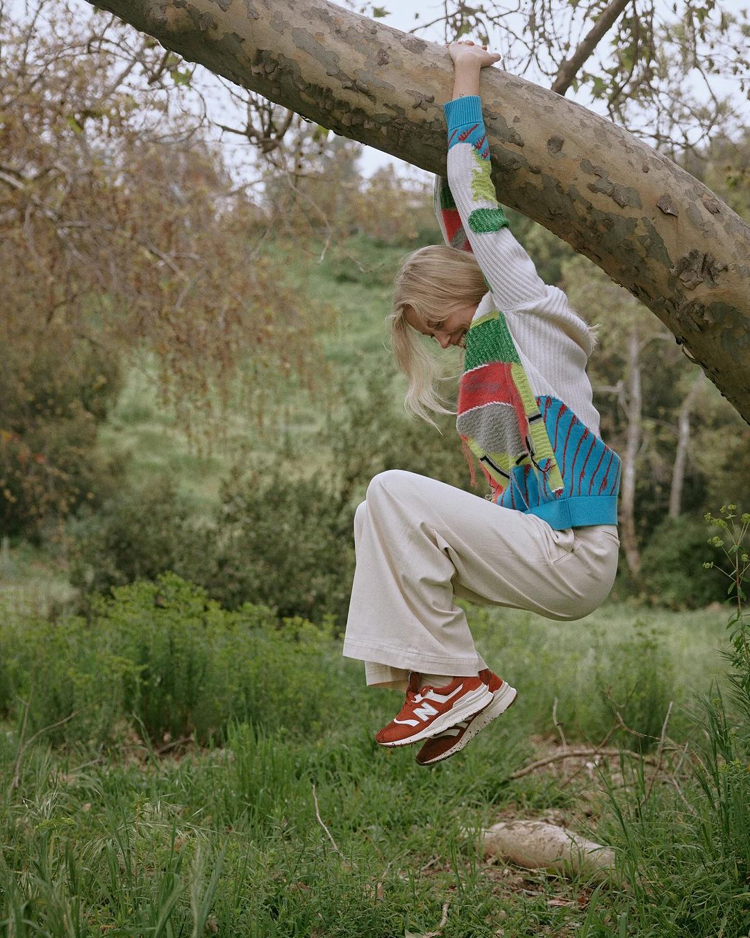 Blonde woman in colorful sweater and cream pants hangs from a tree branch in a grassy, outdoor setting, showcasing an active lifestyle.