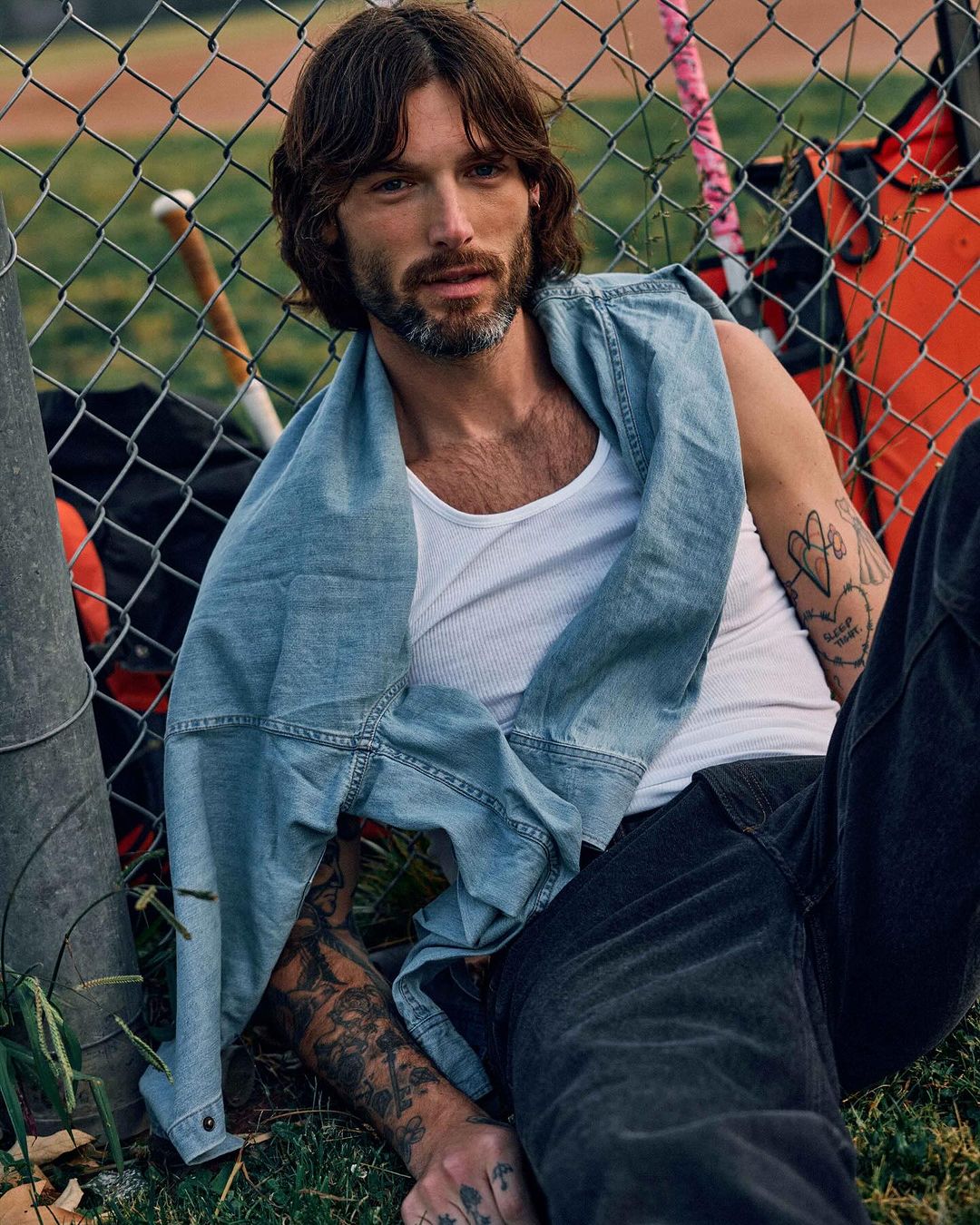 A man with long brown hair is casually leaning against a chain-link fence outdoors, wearing a white tank top and denim jacket.