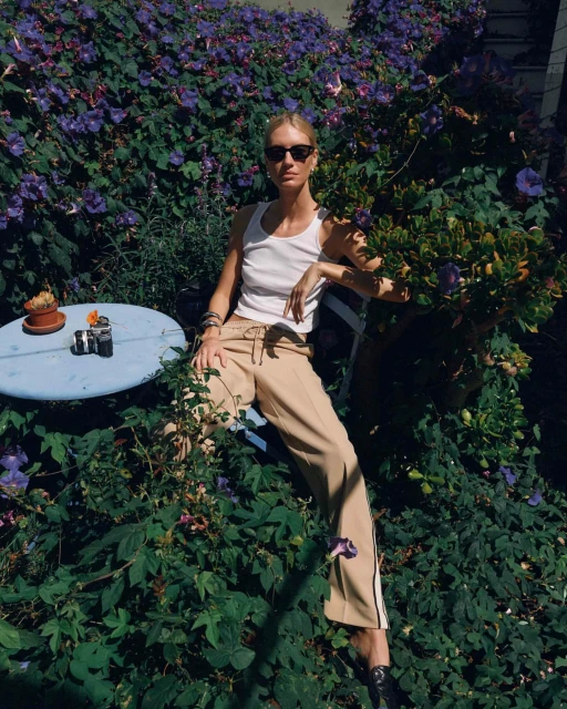 A young adult female model sits in a garden with blue flowers, wearing sunglasses, a white tank top, and tan track pants. She rests her arm.