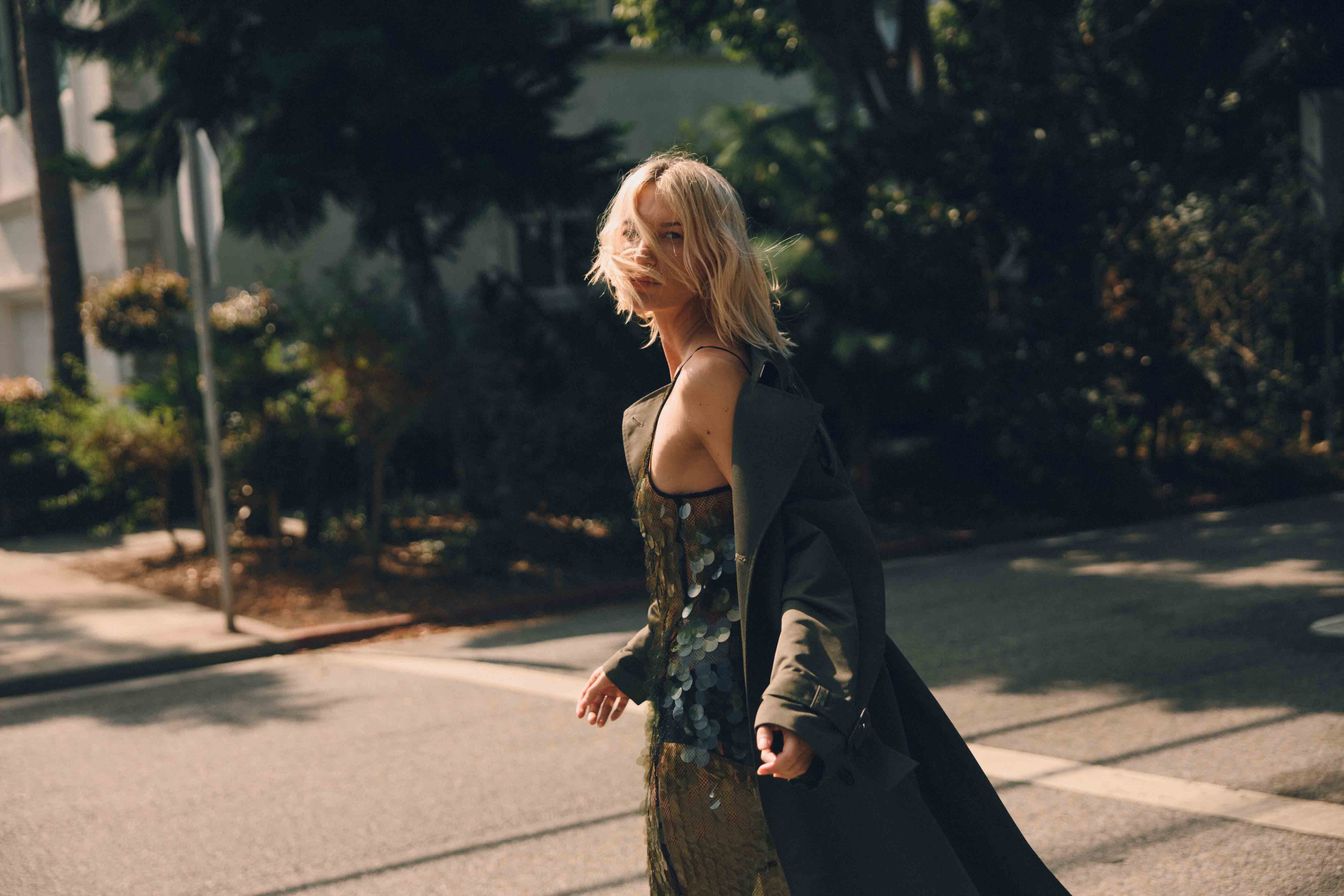 Blonde woman in a sequin dress and trench coat walks across a crosswalk on a sunny city street lined with trees and buildings.