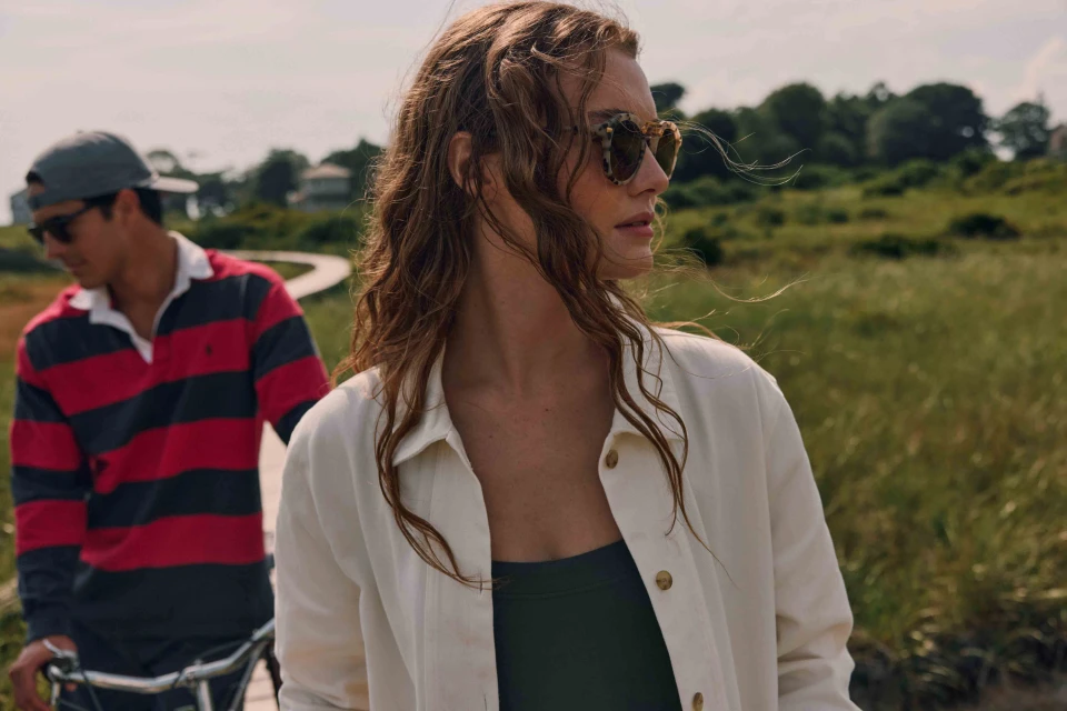 Two people on a grassy path; a woman in sunglasses and a white jacket is in the foreground, and a man walks behind her.