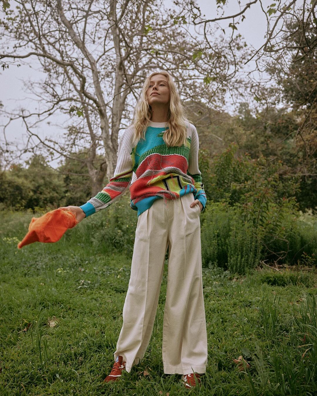Adult woman stands in a grassy field wearing a colorful knit sweater and cream trousers while holding an orange hat in her hand.