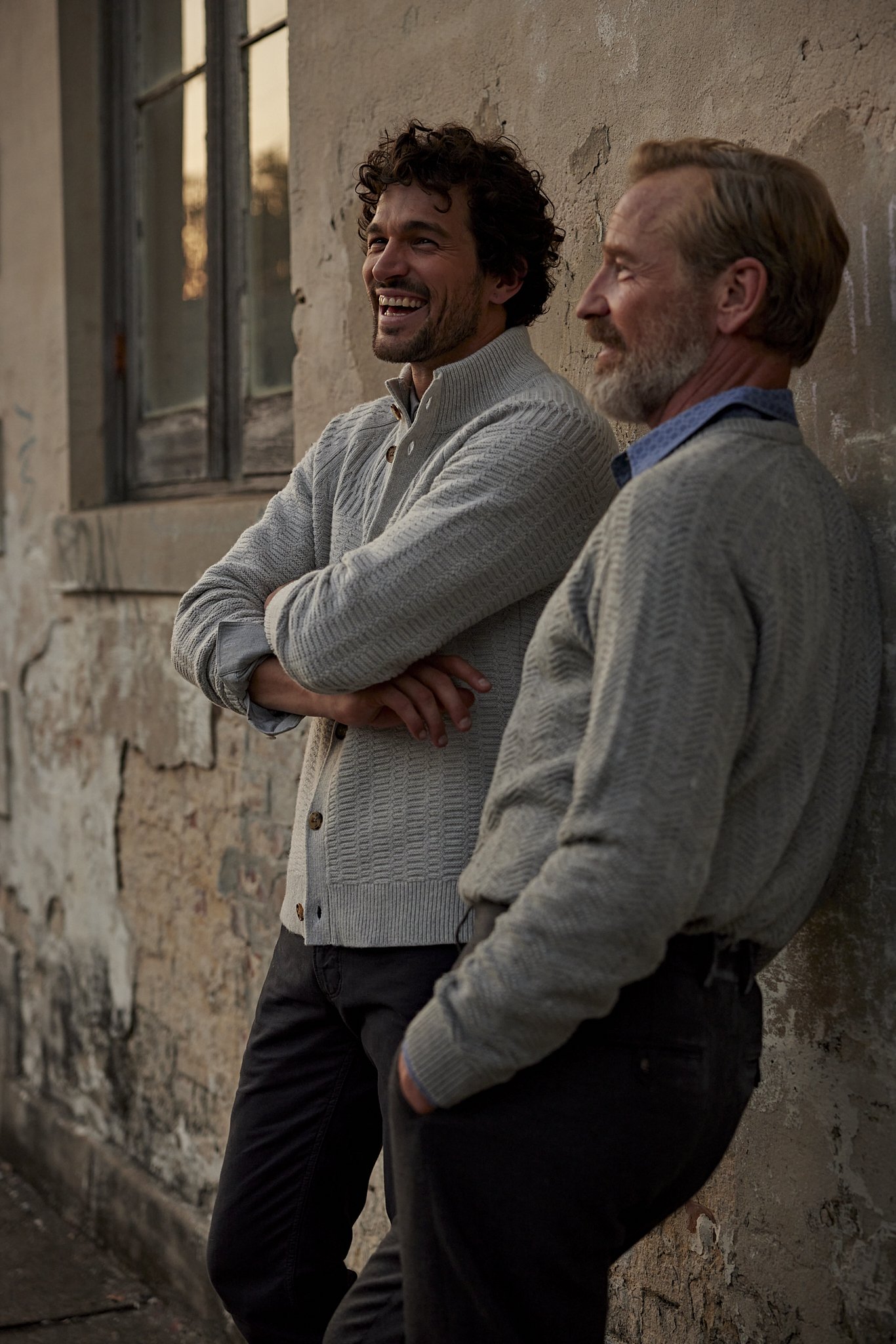 Two men in sweaters lean against a textured wall next to a window during daytime. One man is laughing and looking off camera.