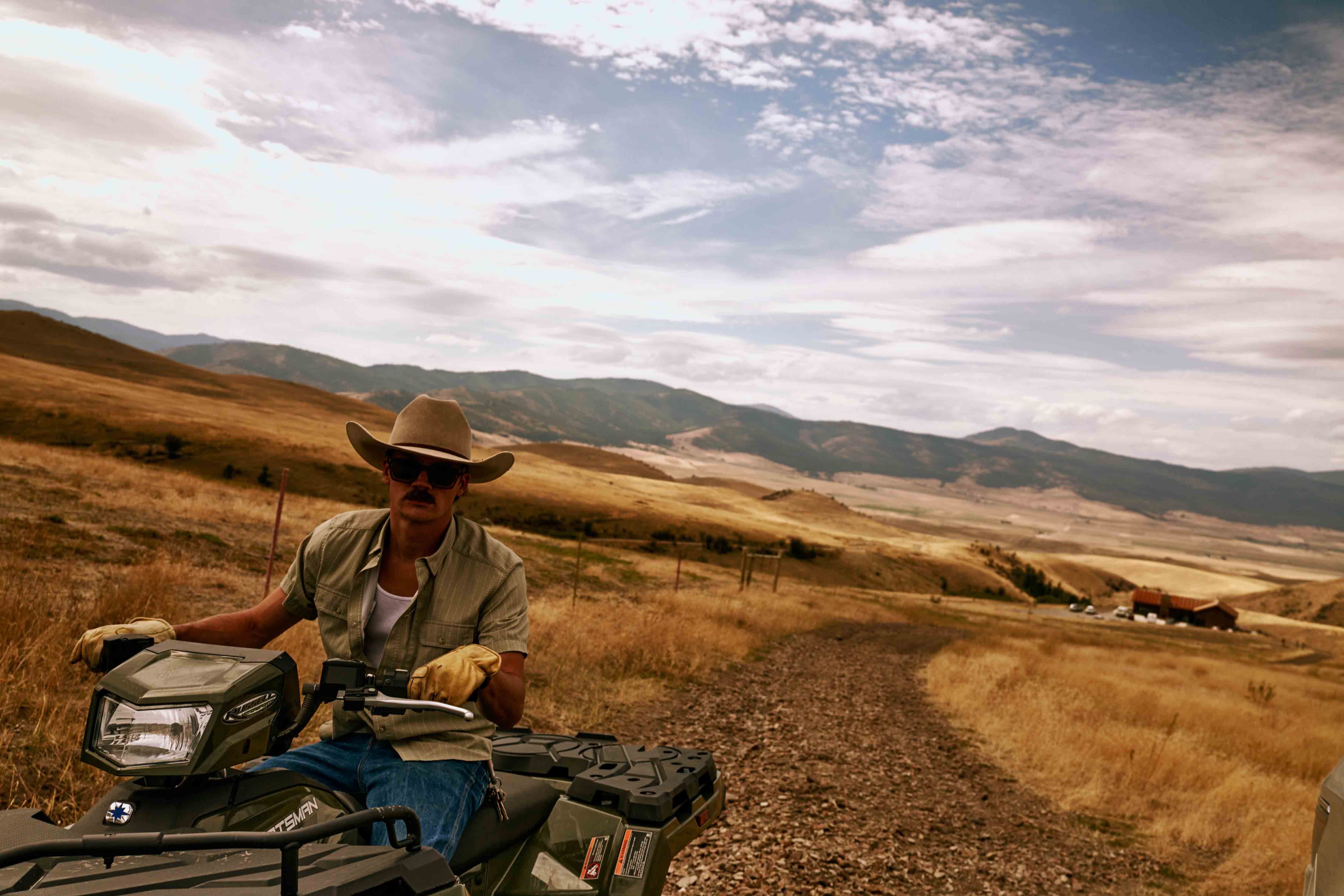 Adult male with a mustache in a cowboy hat on a four-wheeler in a grassy, hilly, outdoor environment with mountains in the background.
